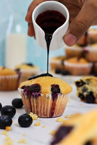 Stock photo showing close-up view of individual homemade blueberry muffin in paper cake case with pile of muffins on marble cake stand beside a bottle of milk, against a light blue background. An unrecognisable person is pouring a small jug of blueberry coulis over the cake.