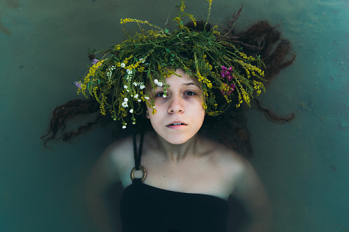 Portrait of a beautiful woman with curly hair enjoying a traditional Slavic summer holiday in the blue lake outdoors, wearing handmade wreath with flowers and grass