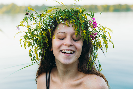 Portrait of a beautiful happy woman with curly hair enjoying a traditional Slavic summer holiday in the blue lake outdoors, wearing handmade wreath with flowers and grass