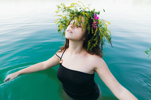 Portrait of a beautiful woman with curly hair enjoying a traditional Slavic summer holiday in the blue lake outdoors, wearing handmade wreath with flowers and grass