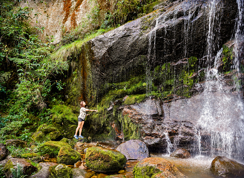 Young woman reaching out to touch a waterfall on a cliff face while out hiking alone in a forest