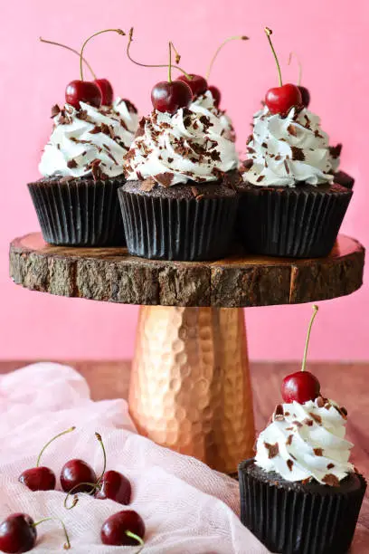 Photo showing a copper and wood cake stand of freshly baked, homemade, Black Forest gateau cupcakes in brown paper cake cases against a pink background. The cup cakes have been decorated with piped whipped cream rosettes topped with morello cherries and sprinkled with chocolate shavings.