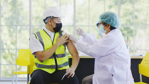COVID-19 Vaccination station in construction industry. Doctor are vaccinating to group engineer worker protect COVID-19 or flu in hospital. People wear face mask protection from virus. health welfare stock photo