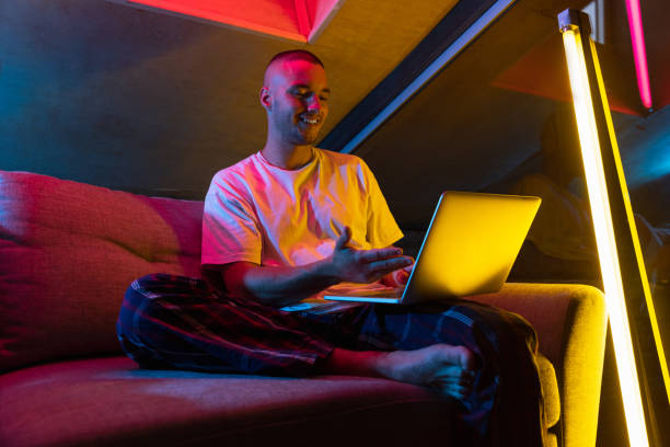 Portrait of handsome young Caucasian man sitting at home in neon orange pink lighted room. Stylish male model indoors. Reflection effect Portrait of handsome young man sitting at home in neon orange pink lighted room. Stylish male model in home clothes indoors. Concept of beauty, fashion, leisure activity, evening and night time cinematic music photos stock pictures, royalty-free photos & images
