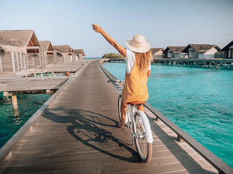 Tropical vacations, young woman with bicycle on wooden pier in the Maldives. Female enjoying bike ride on jetty over coral reef water. Dreamlike destination
