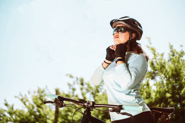 mujer ciclista latinoamericana con ropa deportiva y gafas de sol ajustando su casco protector. concepto de ciclismo y deporte al aire libre. - casco de ciclista fotografías e imágenes de stock