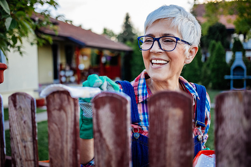 Smiling senior women with grey hair in casual clothes painting fence in her backyard. She using white paint