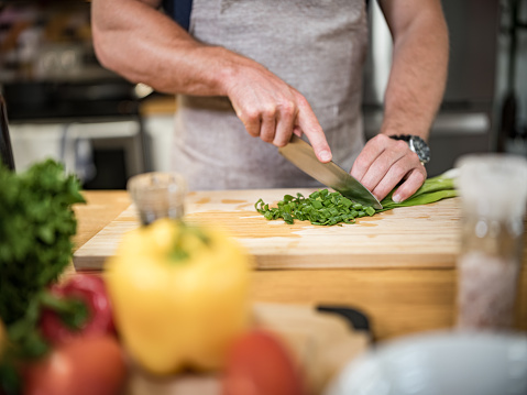 Hands of a Young Caucasian man cutting green onion with kitchen knife in home kitchen. He is wearing casual clothes with chef's apron. He is preparing Italian inspired meal with fresh pasta, meat balls and tossed salad with fresh vegetables and vinaigrette.