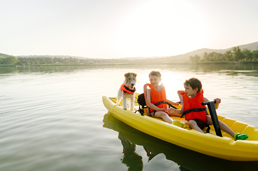 Photo of two cute boys wearing life jackets, and their dog riding a kayak on the lake, on a hot summer day.