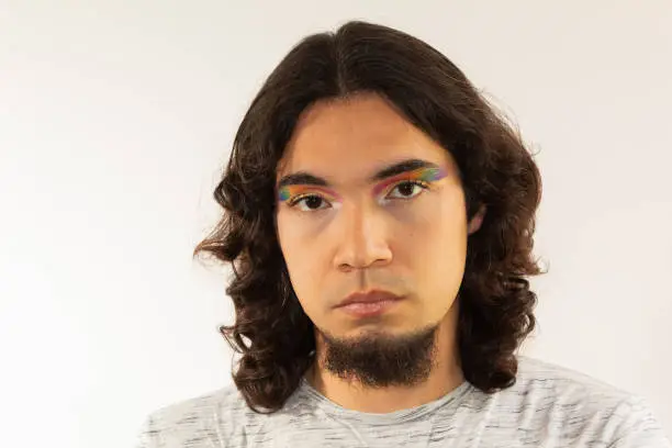 portrait of a gay man made up with a rainbow in his eyes on a white background with a serious face
