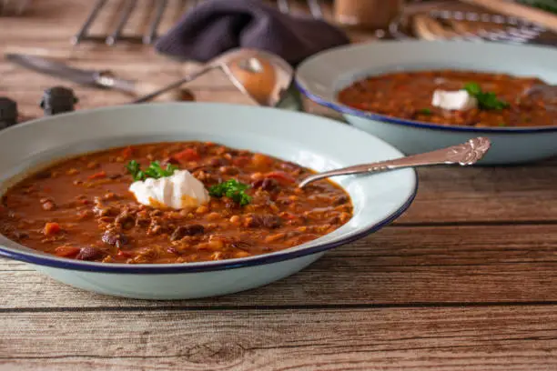 fresh and homemade cooked rustic and spicy bean stew with ground beef, red peppers, chili, onion, garlic and tomatoes served with sour cream on an old fashioned plate with spoon. Closeup view with blurred background