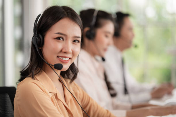 equipe de call center na ásia usa fones de ouvido com microfone. sorria enquanto atende clientes em mesas e computadores. conceito de serviço e consultoria. conceito de comunicação. - neutral look audio - fotografias e filmes do acervo