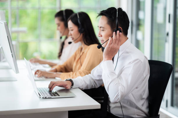 equipe de call center na ásia usa fones de ouvido com microfone. sorria enquanto atende clientes em mesas e computadores. conceito de serviço e consultoria. conceito de comunicação. - neutral look audio - fotografias e filmes do acervo