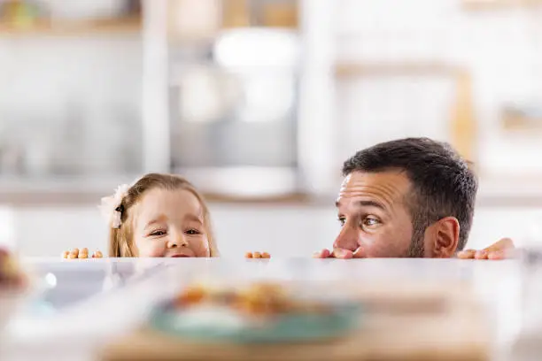 Happy girl and her single father having fun while peeking behind the table at home. Copy space.
