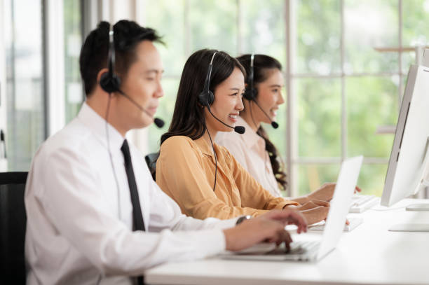 equipe de call center na ásia usa fones de ouvido com microfone. sorria enquanto atende clientes em mesas e computadores. conceito de serviço e consultoria. conceito de comunicação. - neutral look audio - fotografias e filmes do acervo