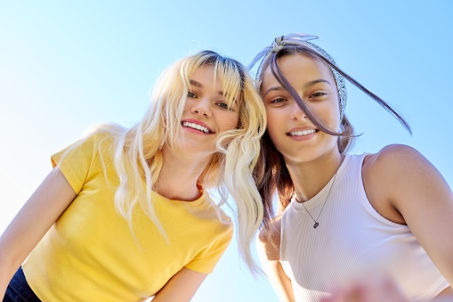 Close-up of happy smiling faces of teenage girls. Two females, blonde and brunette looking at the camera, sky background.