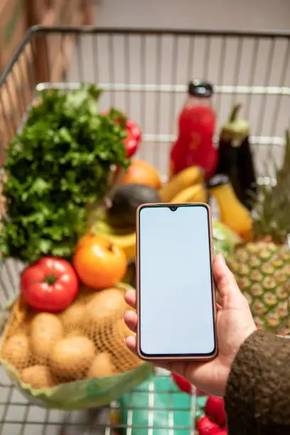 hand holding phone with white screen copy space in grocery shopping cart with fruits and vegetables healthy food