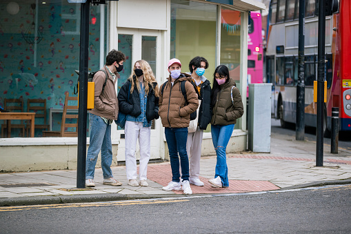 A front-view shot of a group of female and male teenage friends walking through a residential district, they are waiting to cross the road on a footpath in Wallsend, North East England. They are all wearing protective face masks, to try and reduce contact during the COVID-19 pandemic.