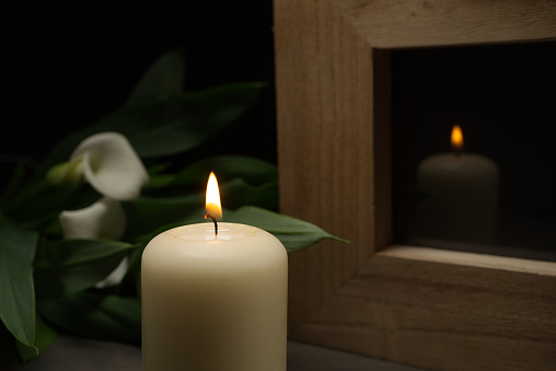 Large white candle burning in the darkness with reflection on glass in a wooden frame and arum lilies illuminated by the candlelight alongside in a conceptual image