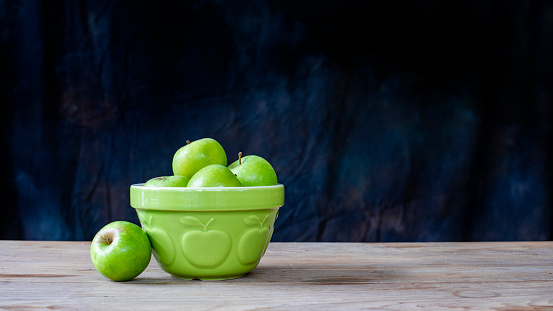 Close up of a bowl of green apples sitting on a table in front of a black background.