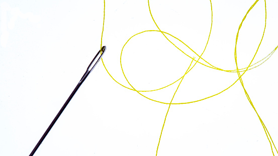 Extreme close up macrophotography of yellow thread threaded through a sewing needle laying against a white background.