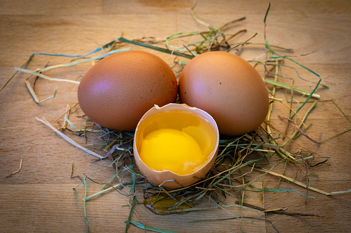 Close up of eggs sitting on a table with straw underneath them. One of the eggs has been broken to be able to see the yolk inside.