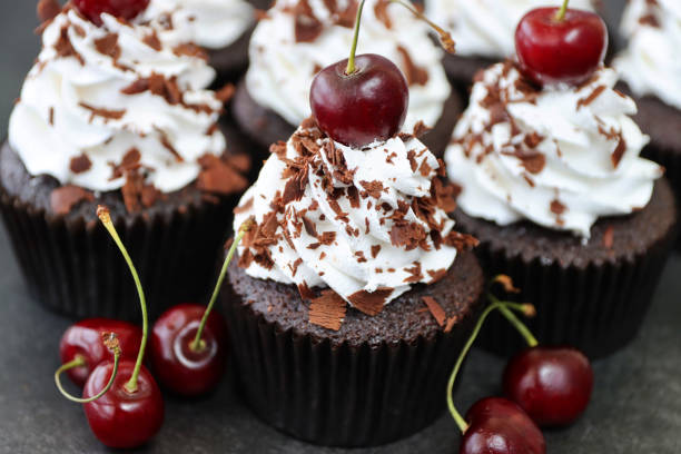 Stock photo showing a batch of freshly baked, homemade, Black Forest gateau cupcakes in brown paper cake cases against a black background. The cup cakes have been decorated with piped whipped cream rosettes topped with morello cherries and sprinkled with chocolate shavings. imagen de primer plano de un lote de cupcakes caseros, gateau de la selva negra en cajas de pasteles de papel marrón, rosetas de crema batida con tuberías cubiertas con cerezas morello espolvoreadas con virutas de chocolate, fondo negro, enfoque en prime