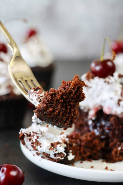 Stock photo showing a homemade, Black Forest gateau cupcake with piped whipped cream rosette topped with morello cherry and sprinkled with chocolate shavings served on a white plate being cut by a metal fork. imagen de corte de tenedor de metal en casera, cupcake gateau de la selva negra sobre plato blanco, roseta de crema batida con tuberías cubierta con cereza morello y espolvoreada con virutas de chocolate, fondo negro moteado, enfoque en primer plano