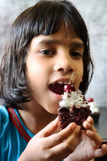 Stock photo showing close-up view of an unwrapped homemade, Black Forest gateau cupcake being eaten by young Indian girl.