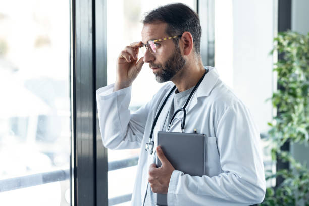 istock Handsome mature male doctor workinh while holding digital tablet looking forward window standing in medical consultation. 1327445168