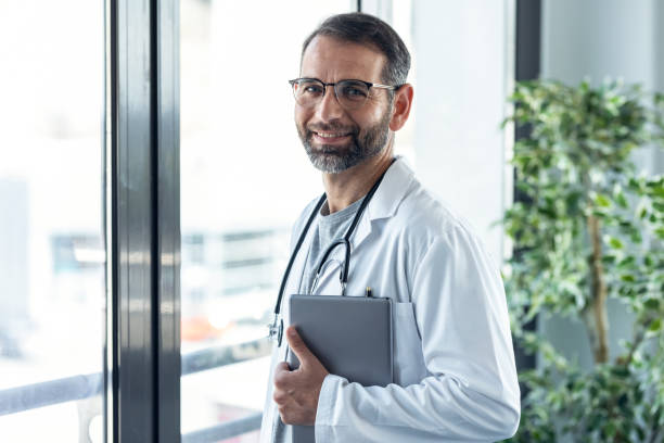 istock Handsome mature male doctor workinh while holding digital tablet looking at camera in the medical consultation. 1327445246
