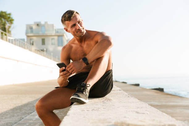Mature sportsman chatting by mobile phone Photo of strong mature sportsman chatting by mobile phone on the beach outdoors. athleticism stock pictures, royalty-free photos & images