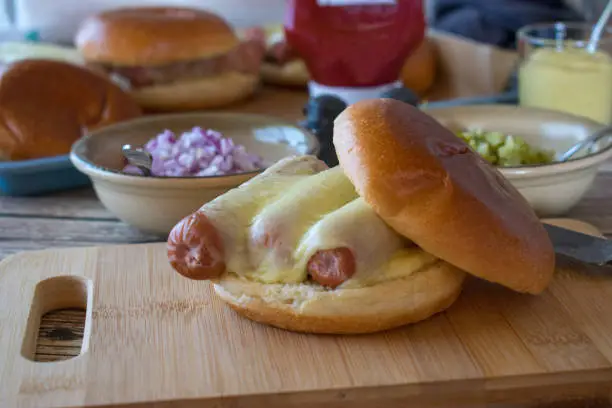 homemade fresh prepared hotdog on a burger bun with melted cheese served with chopped red onions, pickles, mustard and ketchup on a dinner table. Front view with blurred background