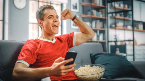 Charismatic Young Adult Man Sitting on a Couch Watches Game on TV, uses Smartphone App for Score, Bet, Statistics, Celebrates Victory when Team Wins Championship. Happy Fan Watches Sport. Charismatic Young Adult Man Sitting on a Couch Watches Game on TV, uses Smartphone App for Score, Bet, Statistics, Celebrates Victory when Team Wins Championship. Happy Fan Watches Sport. checking ice hockey stock pictures, royalty-free photos & images