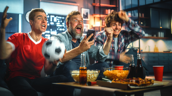 Noche en casa: Tres fanáticos del fútbol sentados en un sofá ven un partido en la televisión, use la aplicación del teléfono inteligente para apostar en línea, celebre la victoria cuando el equipo deportivo gane. Los amigos animan a comer bocadillos photo