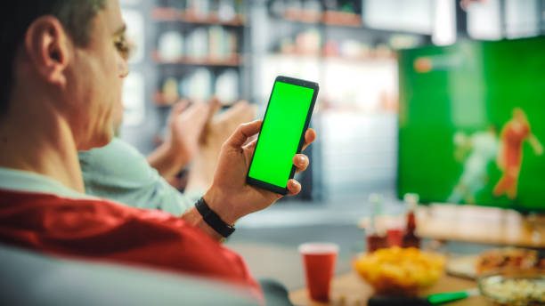 en casa, el fanático del fútbol tiene un teléfono inteligente con pantalla verde chroma key, con amigos que ven el partido de fútbol en la televisión, animan al equipo de deportes de favortis para ganar el campeonato. fútbol club play. sobre el hombr - match sport leisure games sport apps fotografías e imágenes de stock