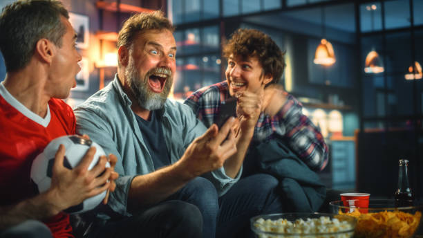 night at home: three soccer fans sitting on a couch watch game on tv, use smartphone app to online bet, celebrate victory when sports team wins. friends cheer eat snacks, watch football play. - gambling imagens e fotografias de stock