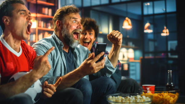 night at home: three soccer fans sitting on a couch watch game on tv, use smartphone app to online bet, celebrate victory when sports team wins. friends cheer eat snacks, watch football play. - gambling imagens e fotografias de stock