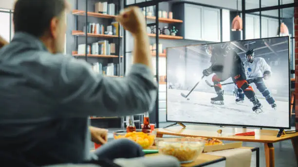 Photo of At Home Ice Hockey Fans Sitting on a Couch Watch Game on TV, Cheer when Favourtite Sports Team to Win the Championship. Screen Shows Professional Players During World Cup. Over the Shoulder
