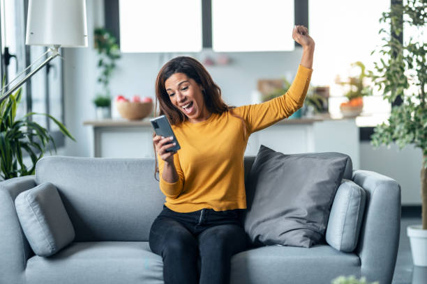 istock Successful young woman using her mobile phone while celebrating something sitting on couch at home. 1327462061