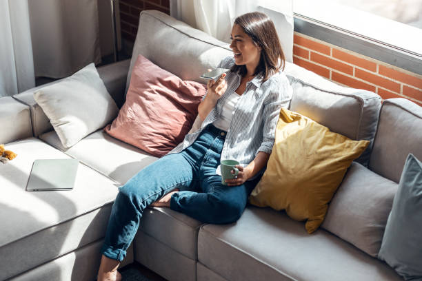 istock Pretty young smiling woman using mobile phone sitting on a couch at home. 1327465846
