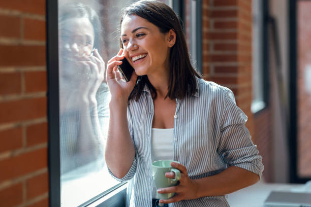 istock Beautiful young woman using her mobile phone while standing next to the window at home. 1327466629