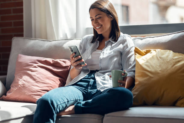 istock Pretty young smiling woman using mobile phone sitting on a couch at home. 1327466679