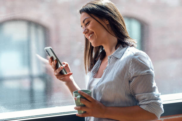 istock Beautiful young woman using her mobile phone while standing next to the window at home. 1327466875
