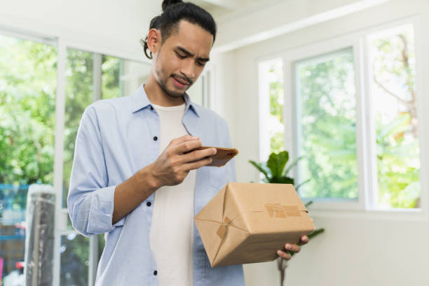 junger asiatischer mann mit smartphone, um ein bild von einem barcode auf einem paket-etikett für die rückgewinnung zu machen - return stock-fotos und bilder