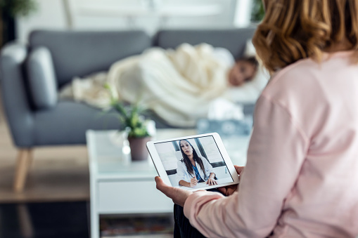 Shot of mature mother making videocall with young female doctor while caring for her sick daughter in living room at home.