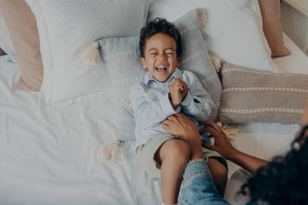 adorable petit fils couché sur des oreillers au lit et riant pendant que maman le chatouille - tickling photos et images de collection
