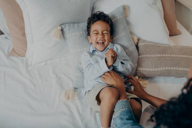 adorable little son lying on pillows in bed and laughing while mom tickling him - tickling imagens e fotografias de stock