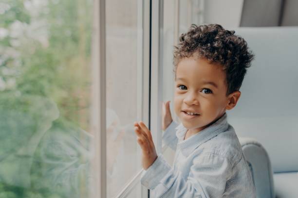 istock Cute mixed race little curly haired boy in light blue colored shirt next to large window 1327485568