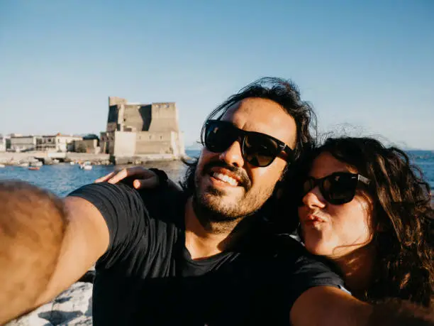 Pov view of a couple taking a selfie near Castel dell'Ovo in Naples, Italy. They are looking at camera, smiling.